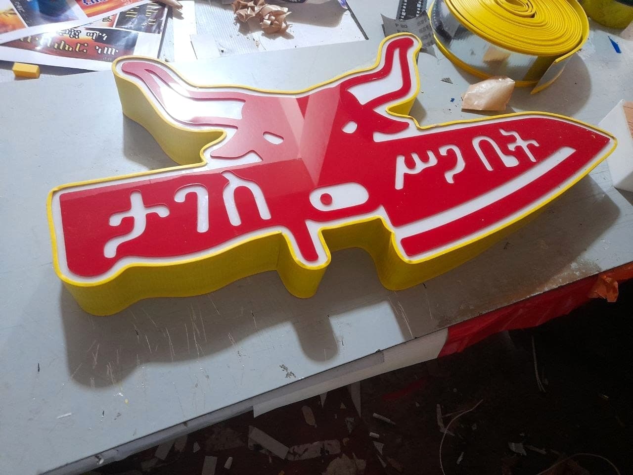 Contour-cut sign in red, white and yellow acrylic with raised Amharic lettering, cut to the outline of the artwork and resting on the bench