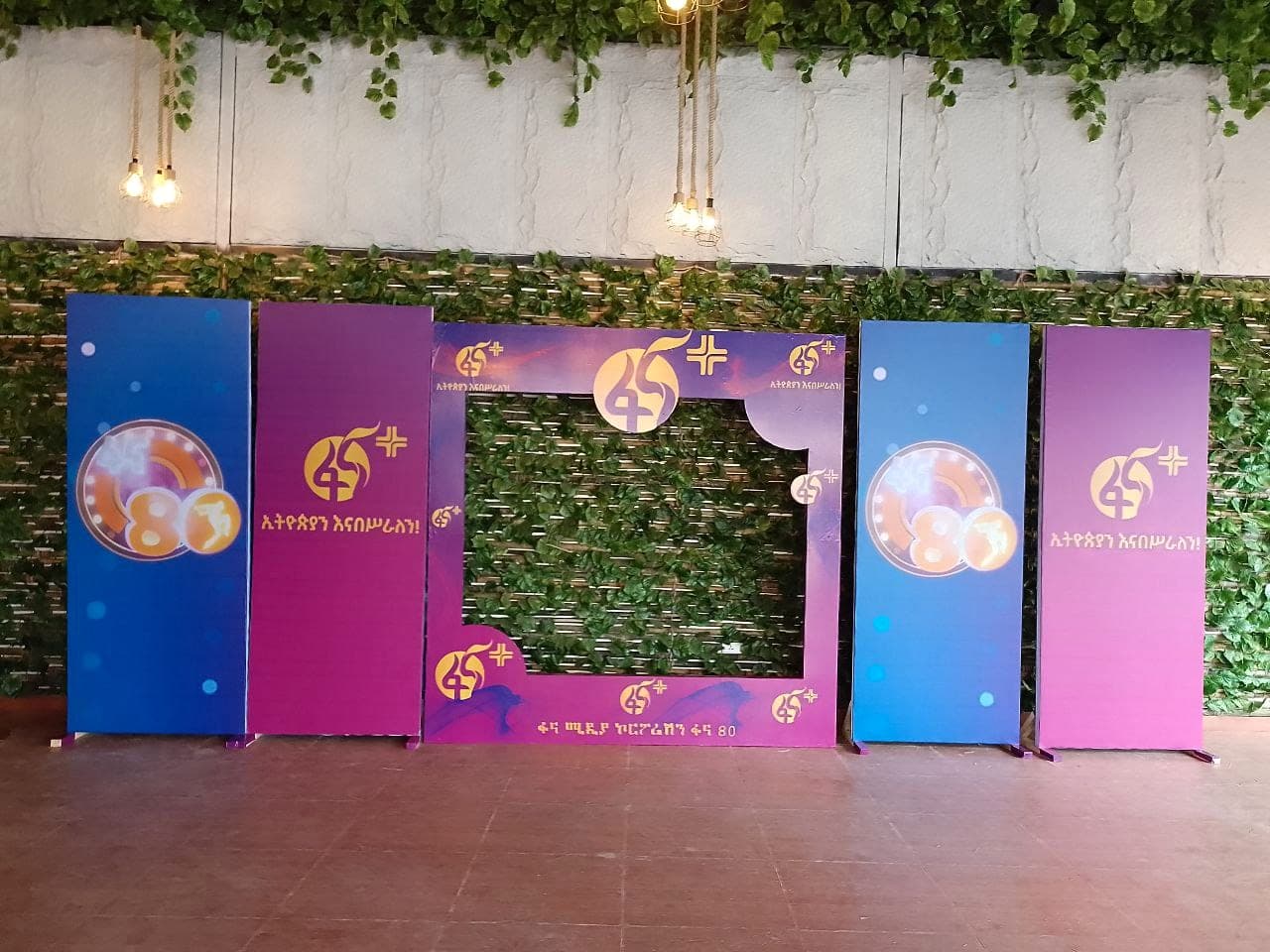 Set of blue and magenta Fana 45 pillar banners and a cut-out photo frame arranged against a foliage wall