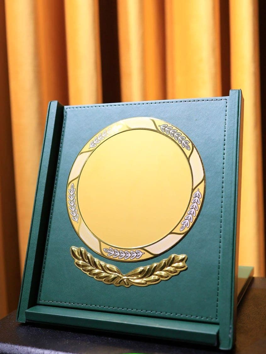 Green leatherette presentation plaque with a blank gold medallion and laurel spray, standing open on a stand