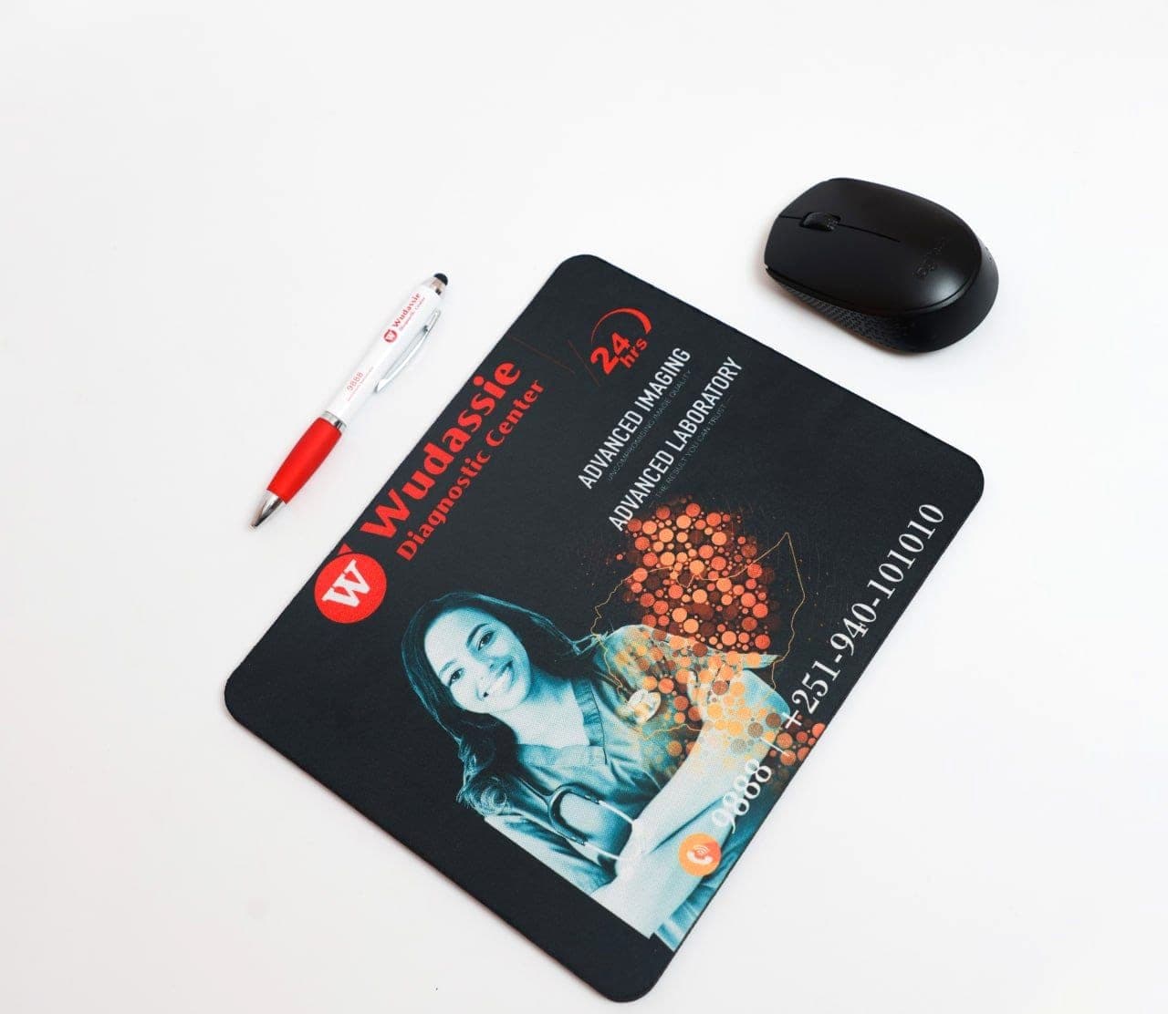 Black printed mousepad for Wudassie Diagnostic Center beside a matching branded pen and a wireless mouse
